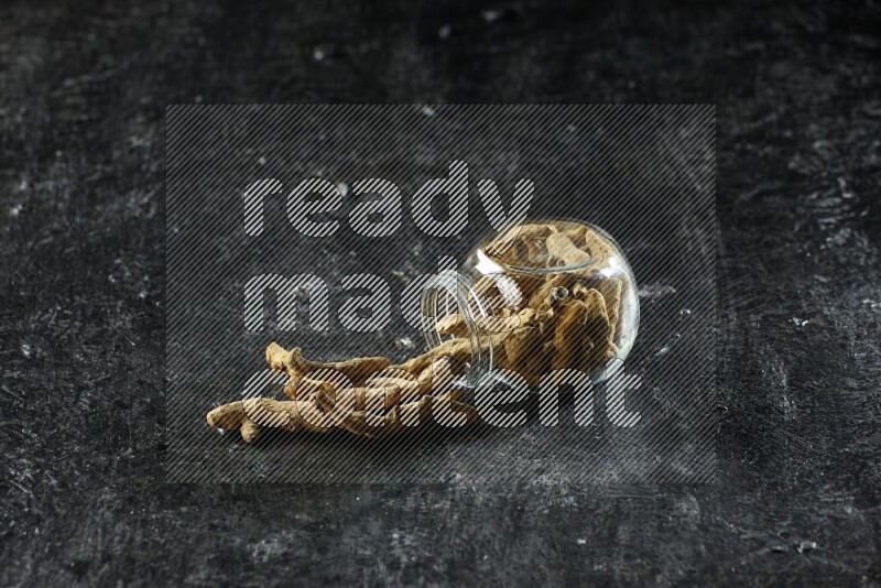 A flipped glass spice jar full of dried turmeric whole fingers and fingers fell out of it on textured black flooring