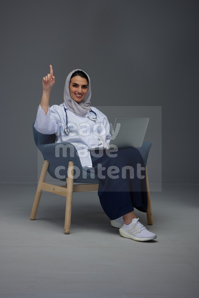 A doctor wearing a light gray head scarf setting on a dark grey chair and using a phone eye level on a grey background