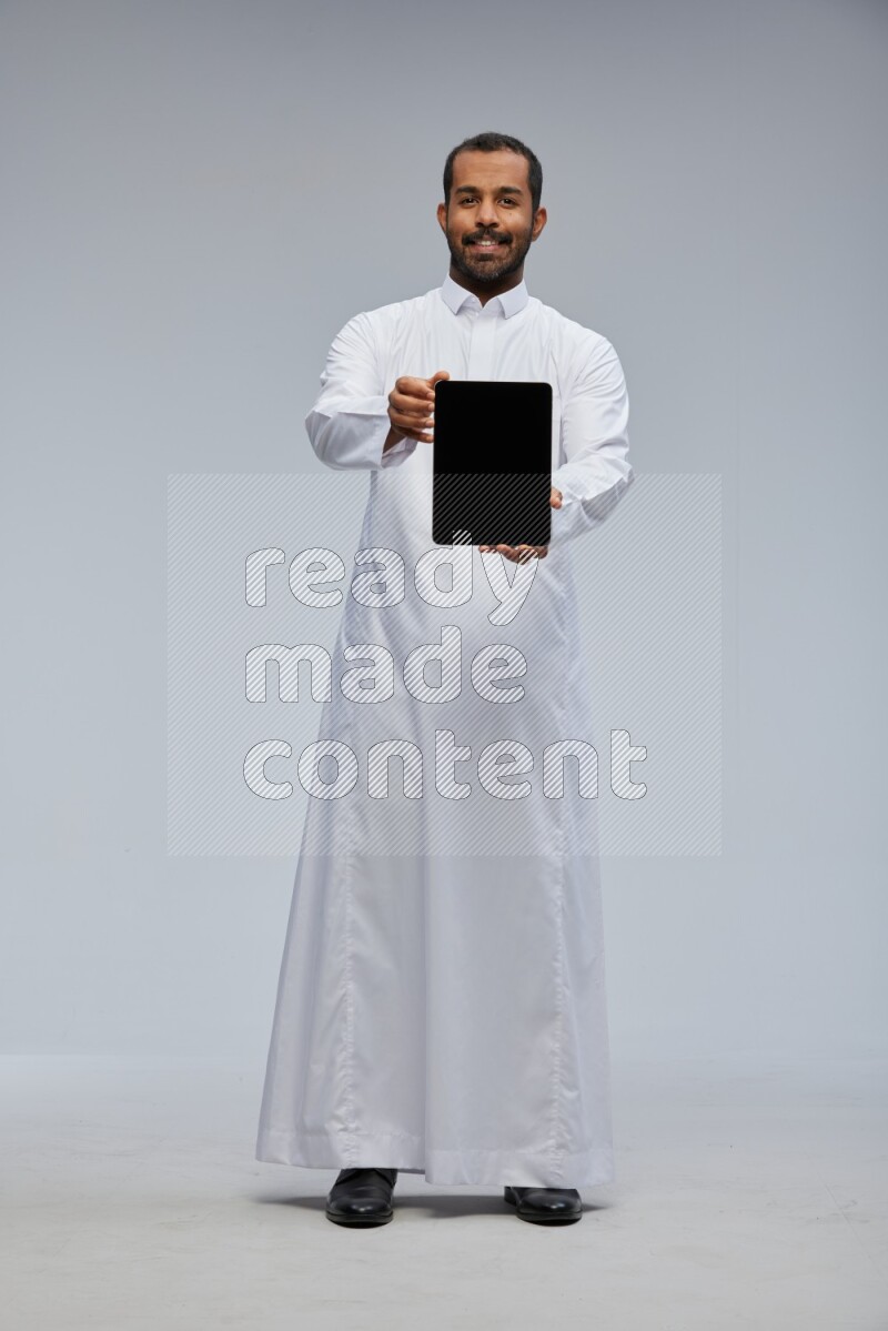 Saudi man wearing Thob standing showing tablet to camera on Gray background