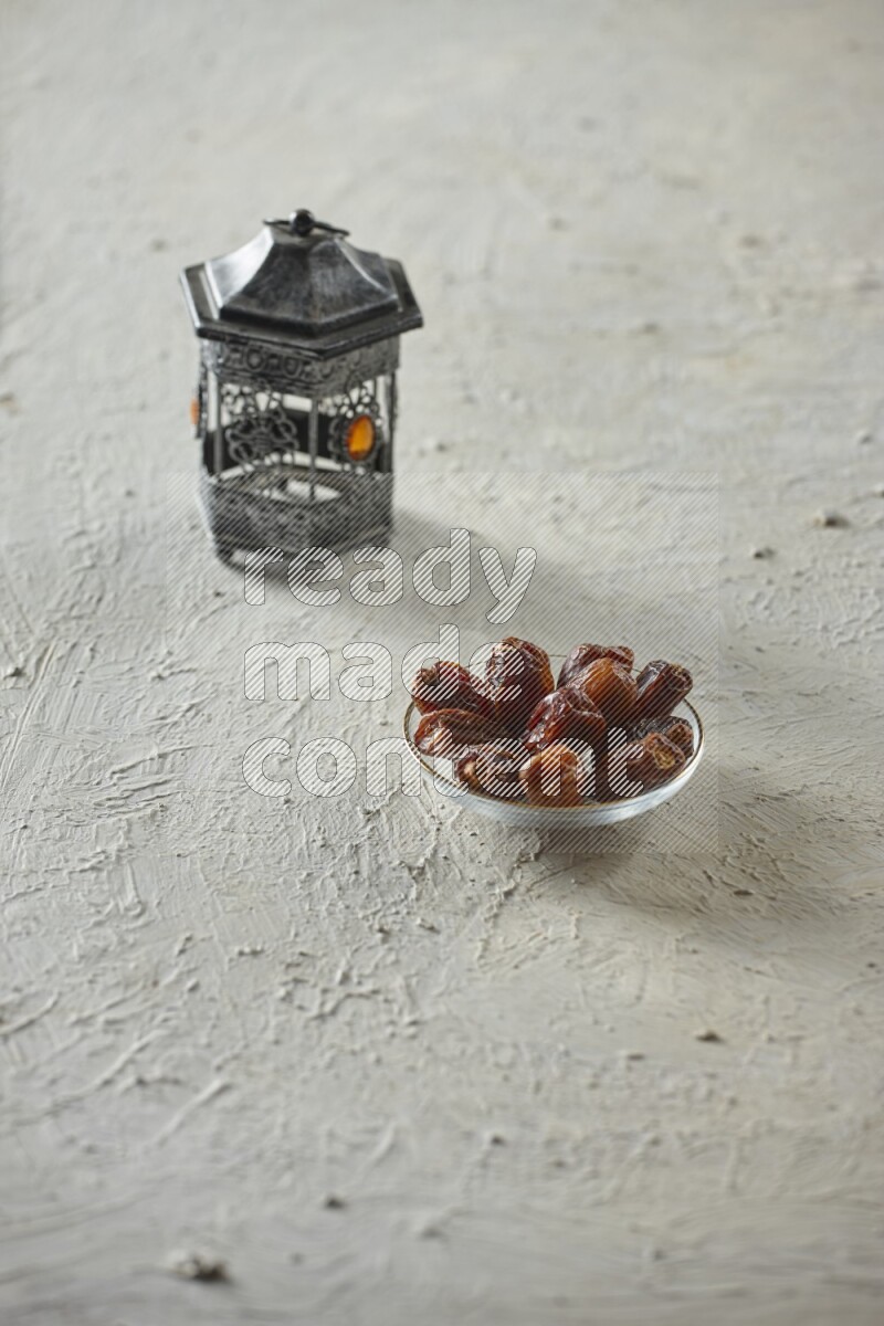 A silver lantern with different drinks, dates, nuts, prayer beads and quran on textured white background