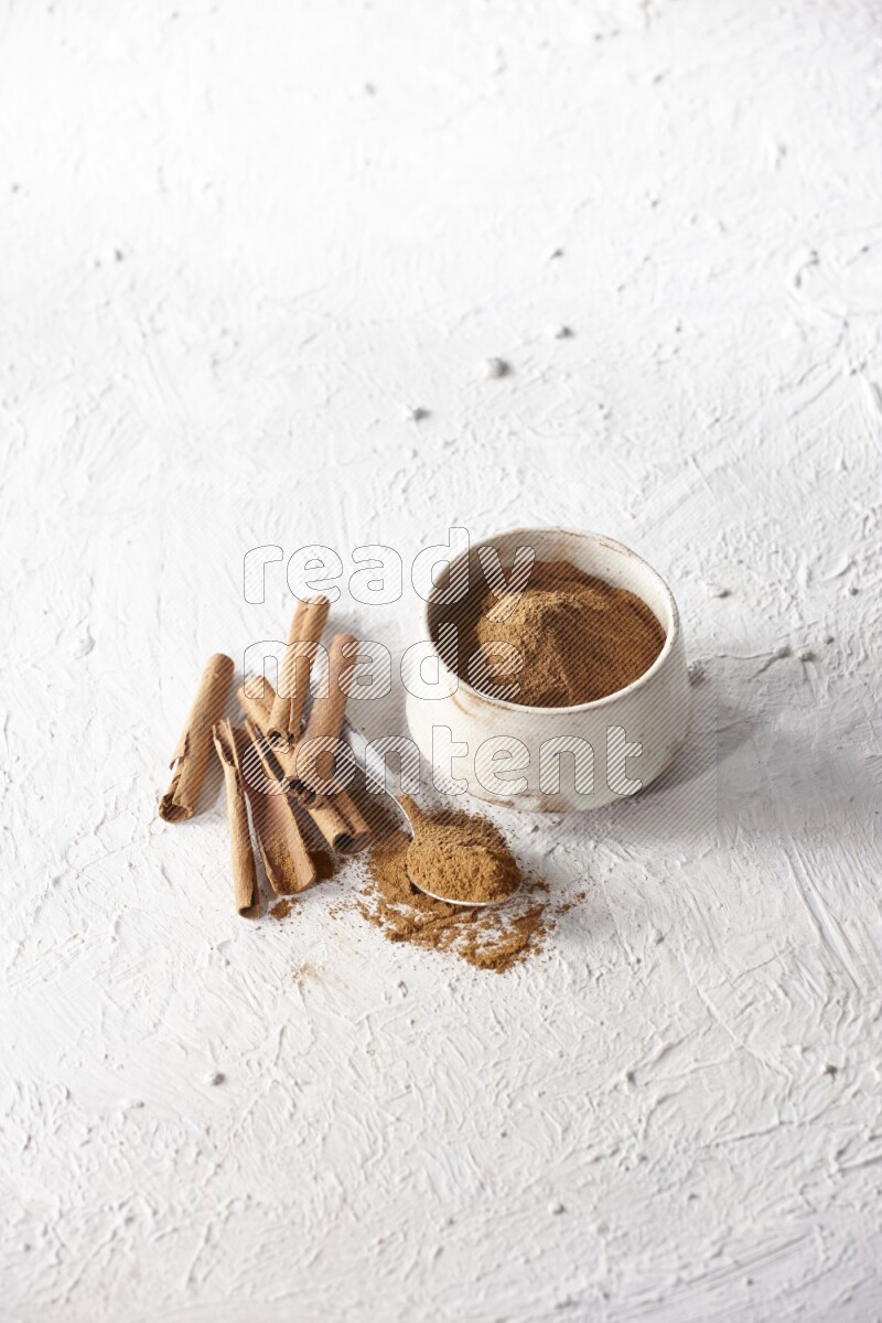 Ceramic beige bowl full of cinnamon powder and a metal spoon with cinnamon sticks next of it on a textured white background