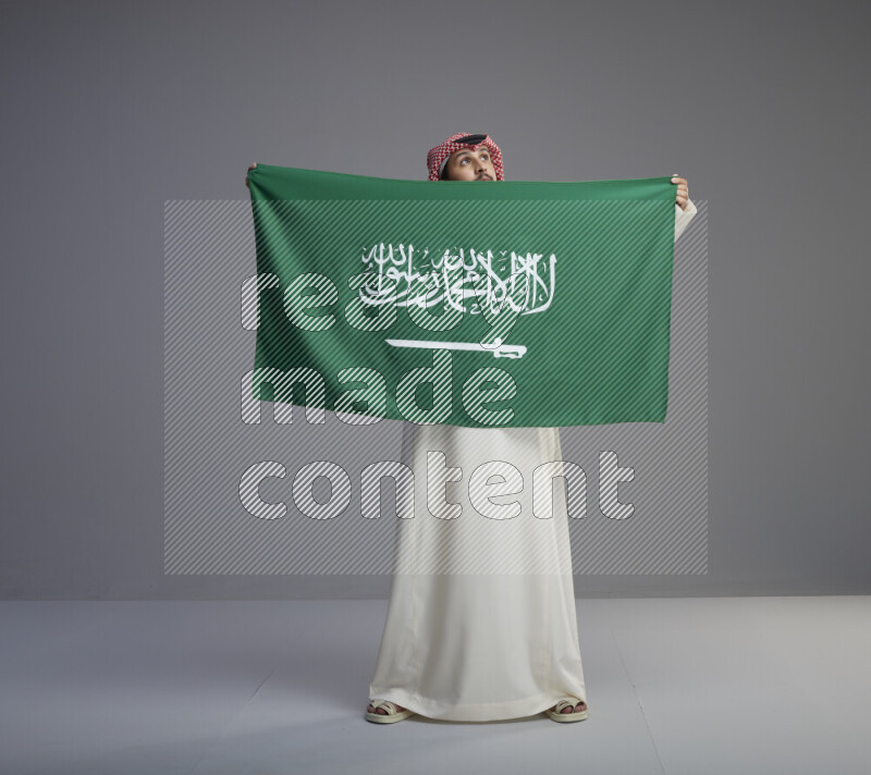 A saudi man standing wearing thob and red shomag holding big saudi flag on gray background