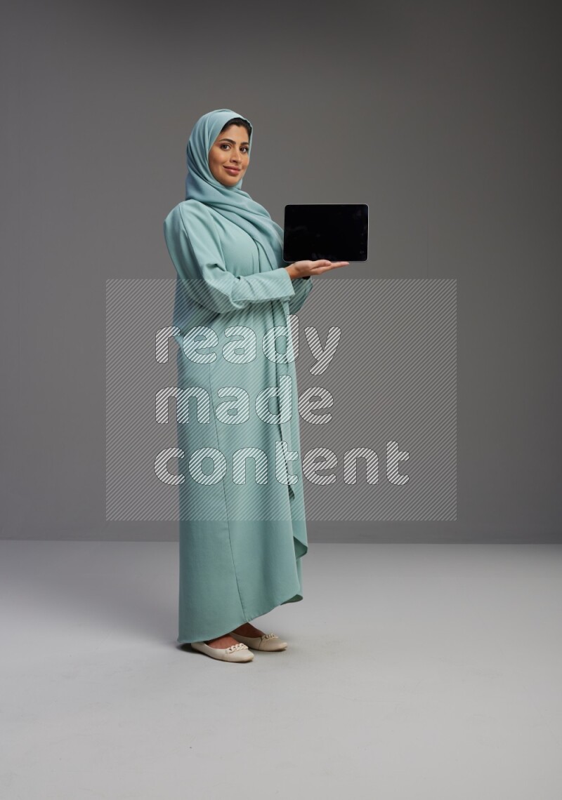 Saudi Woman wearing Abaya standing showing tablet to camera on Gray background