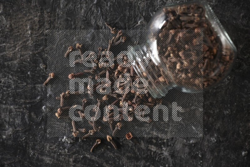 A flipped glass spice jar full of cloves and cloves came out of it on textured black flooring