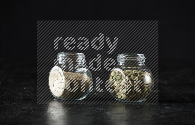 2 glass spice jars full of cardamom powder and cardamom seeds on textured black flooring