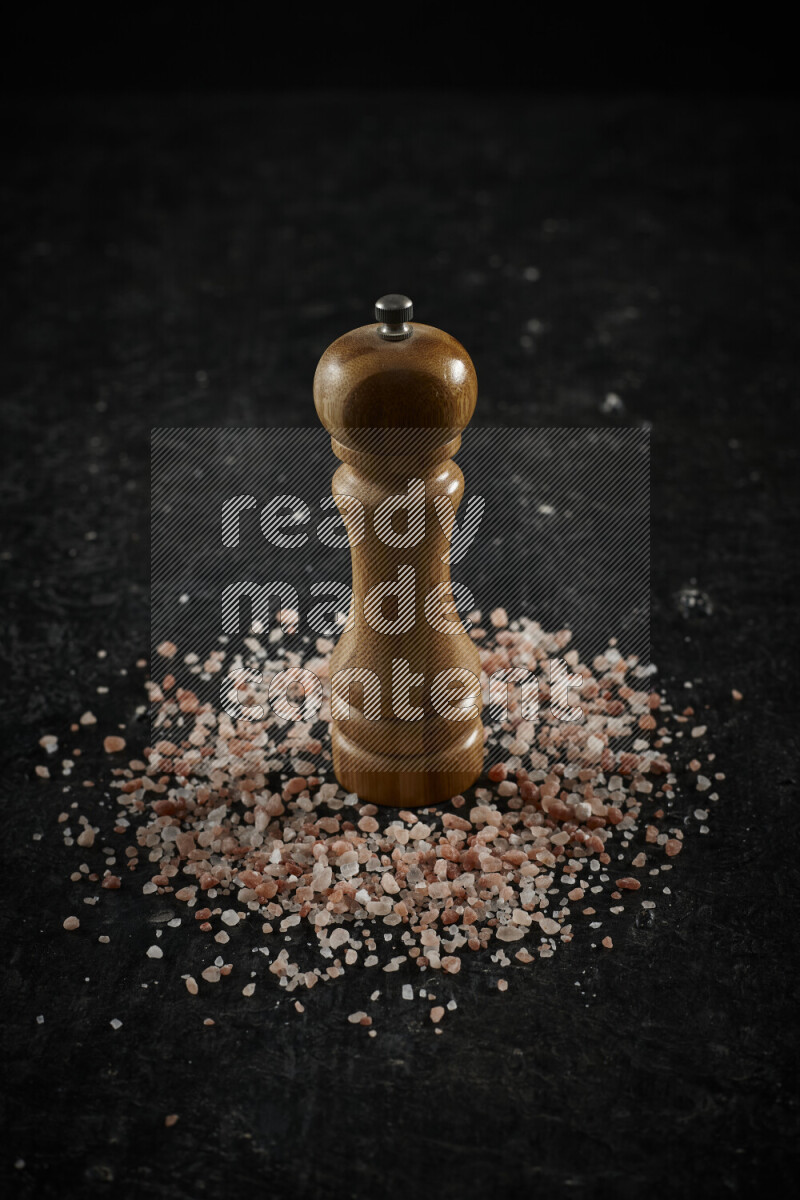 A wooden grinder standing upright and surrounded by coarse pink himalayan salt on black background