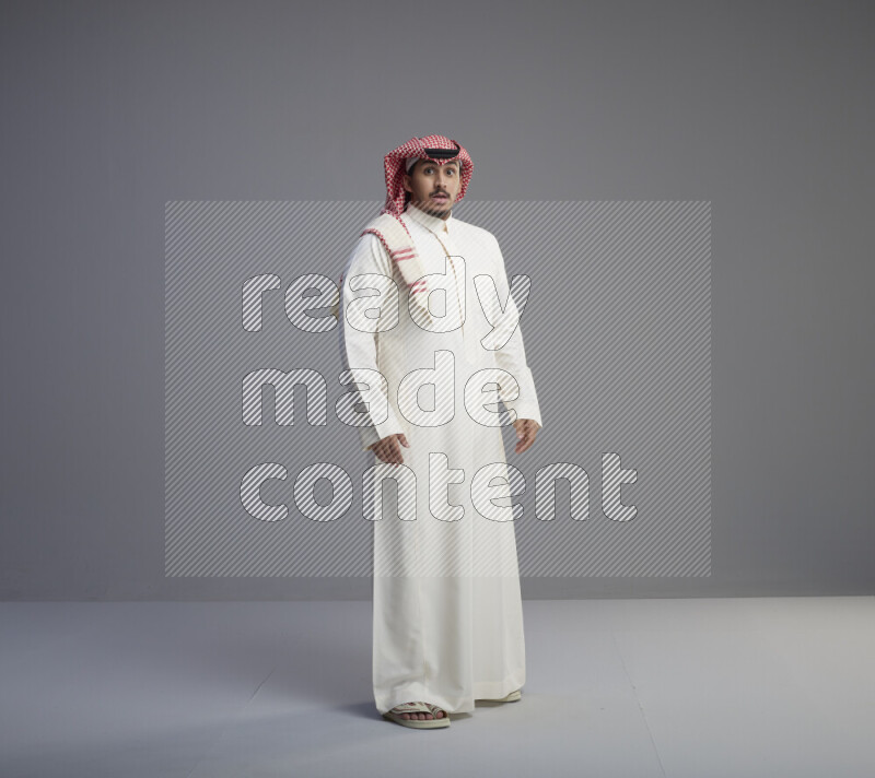 A Saudi man standing wearing thob and red shomag interacting with the camera on gray background