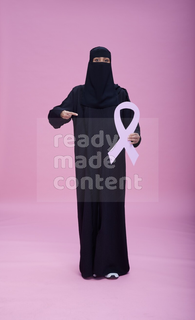 Saudi woman wearing abaya and niqab standing holding awareness ribbon on pink background