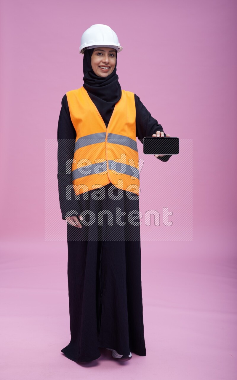 Saudi woman wearing Abaya with engineer vest and helmet standing showing phone to camera on pink background