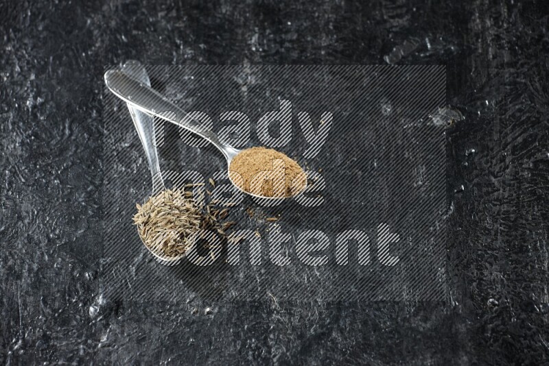 2 metal spoons full of cumin seeds and cumin powder on a textured black flooring