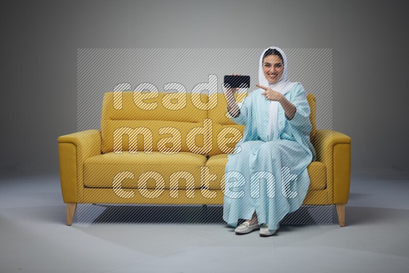 A Saudi woman wearing a light blue Abaya and white head scarf sitting on a yellow sofa and showing her phone's screen vertically and horizontally eye level on a grey background