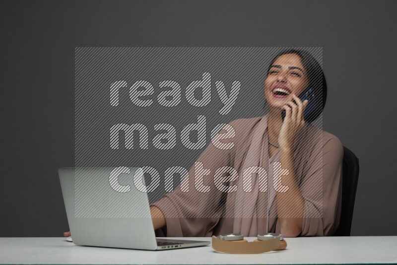 A Saudi woman Sitting on her desk Calling  on a Gray Background wearing Brown Abaya