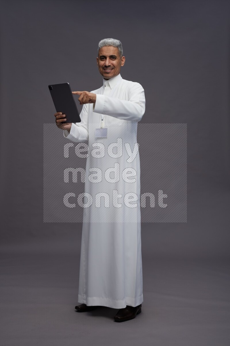 Saudi man wearing thob with neck strap employee badge standing working on tablet on gray background