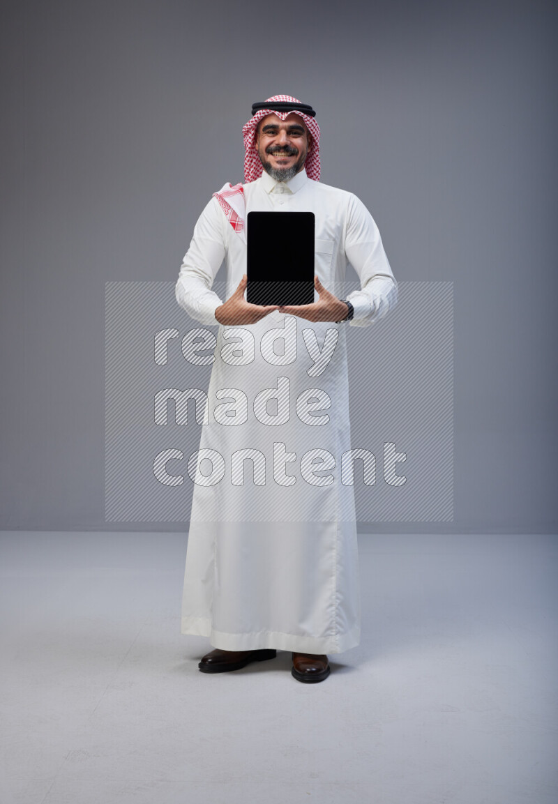 Saudi man Wearing Thob and red Shomag standing showing tablet to camera on Gray background