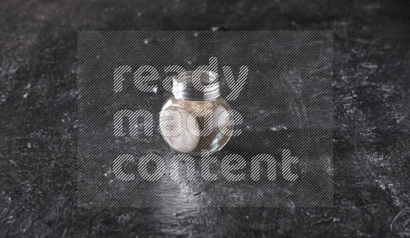 A glass spice jar full of black pepper powder on textured black flooring