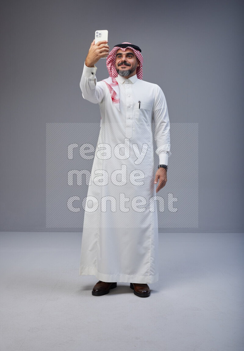 Saudi man Wearing Thob and red Shomag standing taking selfie on Gray background