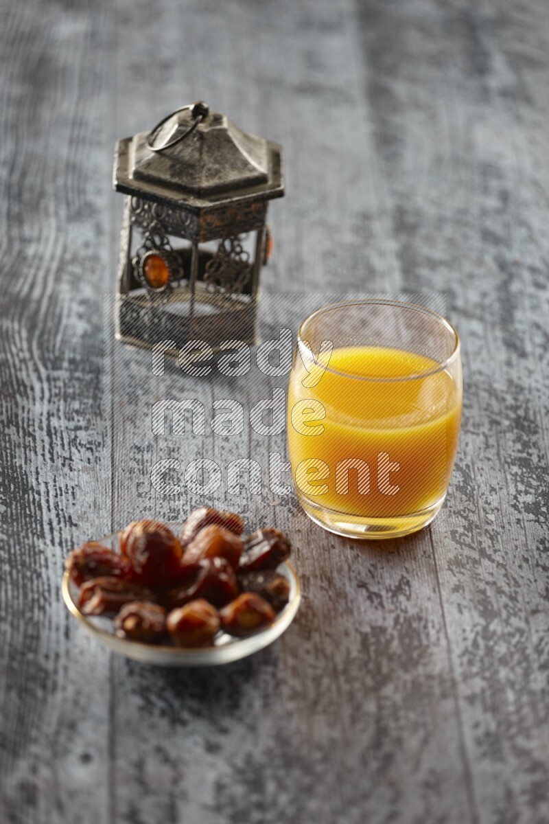 A silver lantern with different drinks, dates, nuts, prayer beads and quran on grey wooden background
