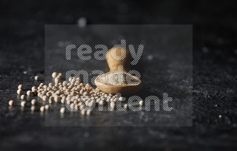 A wooden spoon full of white pepper powder with white pepper beads on textured black flooring