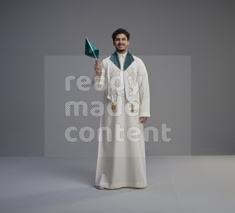A Saudi man standing wearing thob and saudi flag scarf and holding small saudi flag on gray background