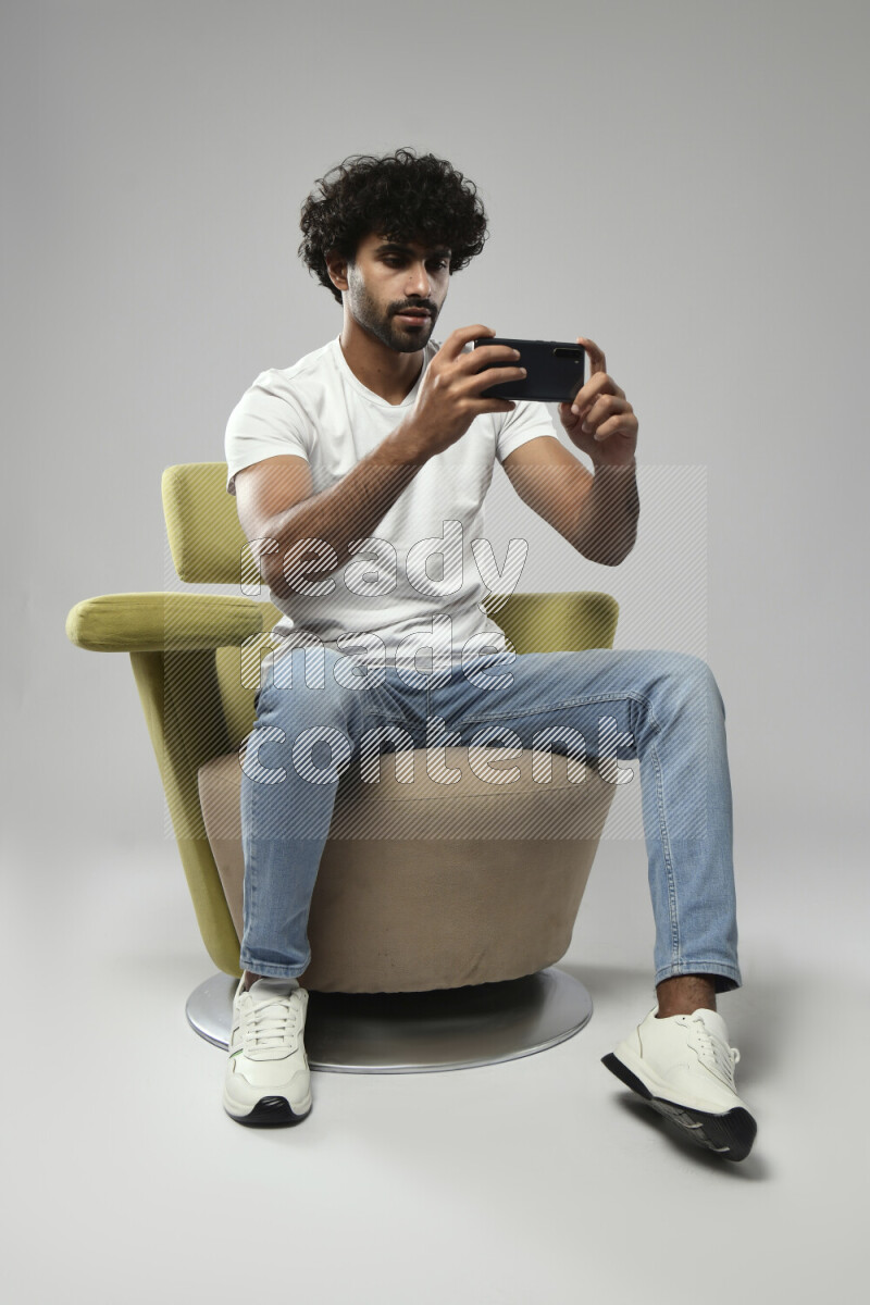 A man wearing casual sitting on a chair shooting with his phone on white background