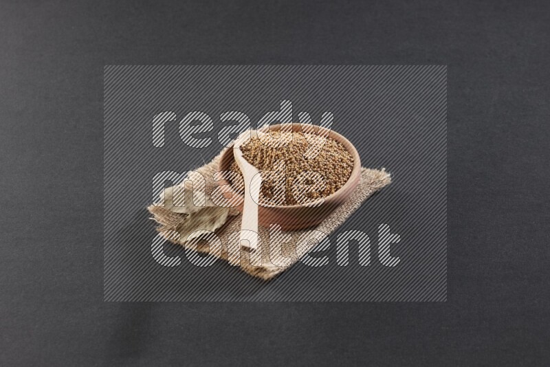 A wooden bowl and spoon full of mustard seeds on a piece of burlap on a black flooring