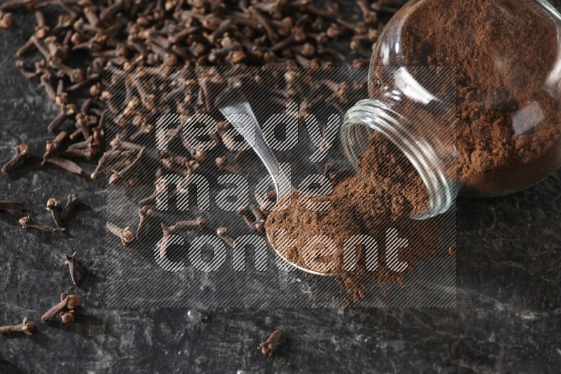 A flipped glass spice jar and a metal spoon full of cloves powder with cloves spread on textured black flooring