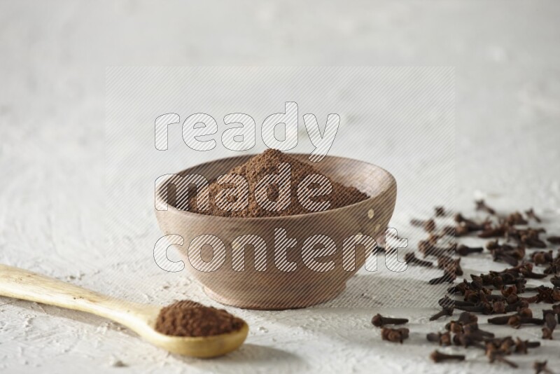 A wooden bowl and wooden spoon full of cloves powder with cloves spread on textured white flooring