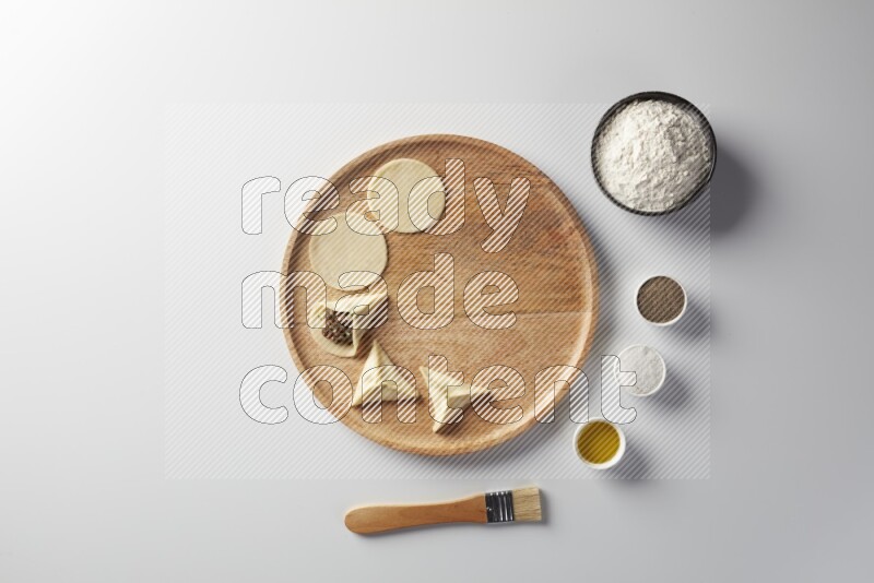 two closed sambosas and one open sambosa filled with meat while flour, salt, black pepper and oil with oil brush aside in a wooden dish on a white background