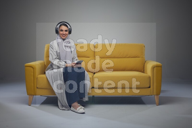 A Saudi woman wearing a light gray Abaya and head scarf sitting on a yellow sofa and using her phone while wearing headphones eye level on a grey background