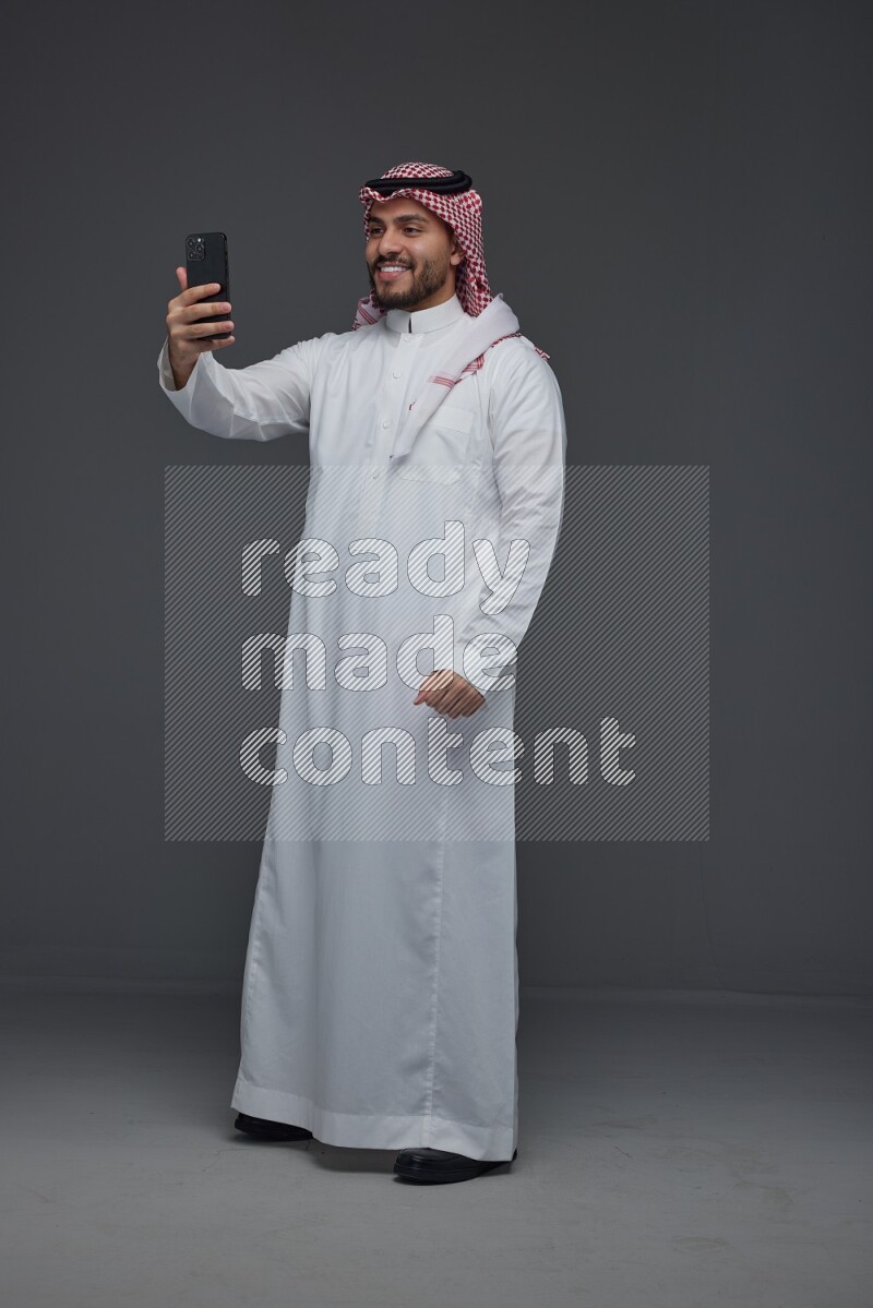 A Saudi man wearing Thobe and Shamgh taking selfie with his phone eye level on a gray background