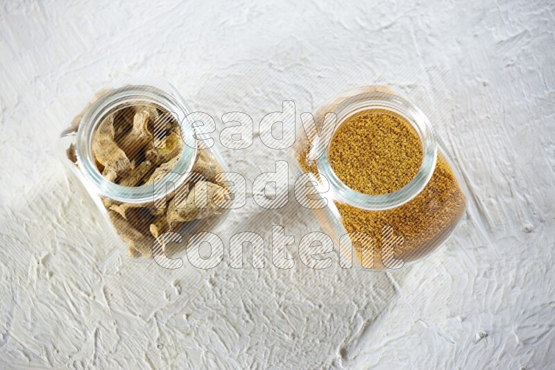 2 glass spice jars full of turmeric powder and dried whole fingers on textured white flooring