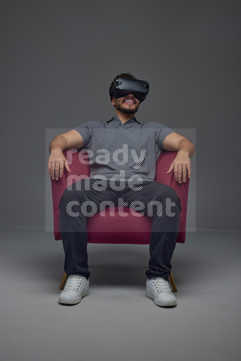 A man wearing casual and wearing VR while sitting on a burgundy chair eye level on a gray background