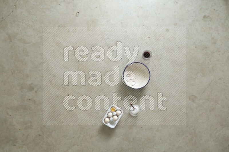 A ceramic bowl beside a carton of eggs and glass bowl full of sugar, vanilla extract, flour on beige marble background