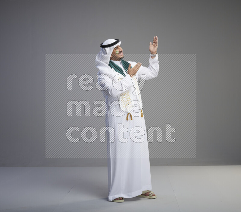 A Saudi man standing wearing thob and white shomag with face painting and Saudi flag scarf on gray background