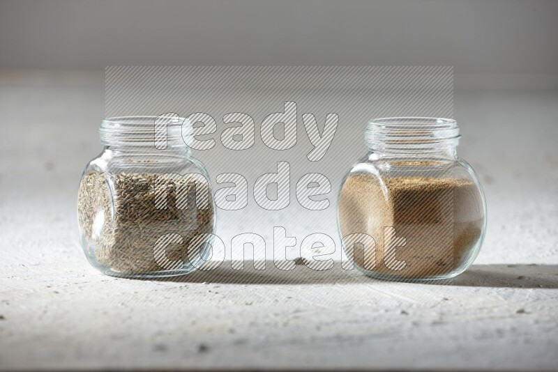 2 glass spice jars full of cumin powder and cumin seeds on textured white flooring