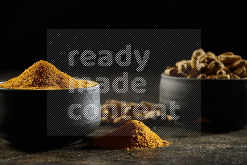 2 pottery black bowls, one full of turmeric powder and the other full of dried turmeric whole fingers on a textured black flooring