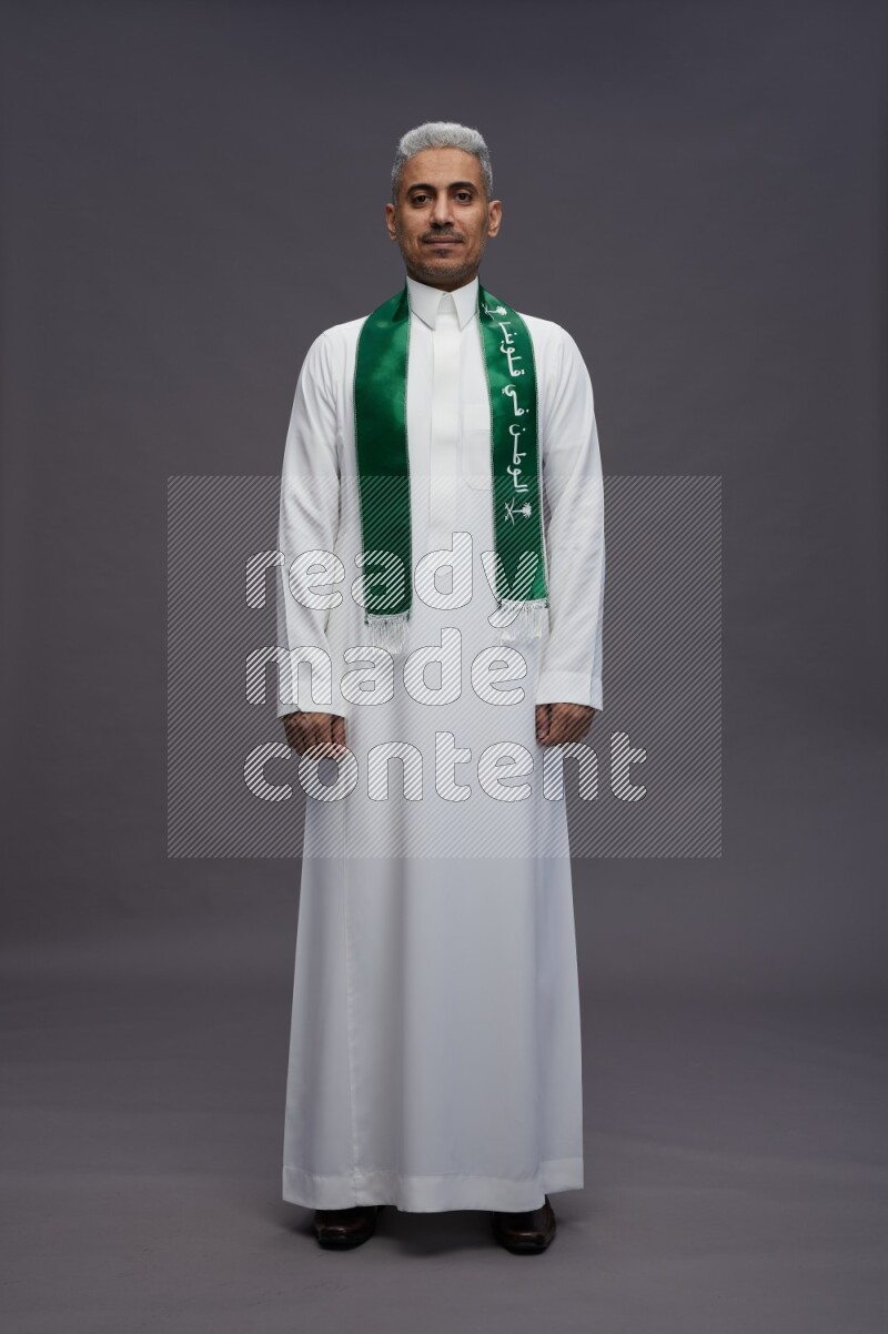 Saudi man wearing thob standing holding Saudi flag on gray background