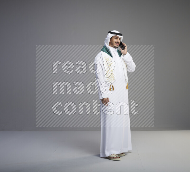 A saudi man standing wearing thob and white shomag with flag scarf on his neck talking on phone on gray background