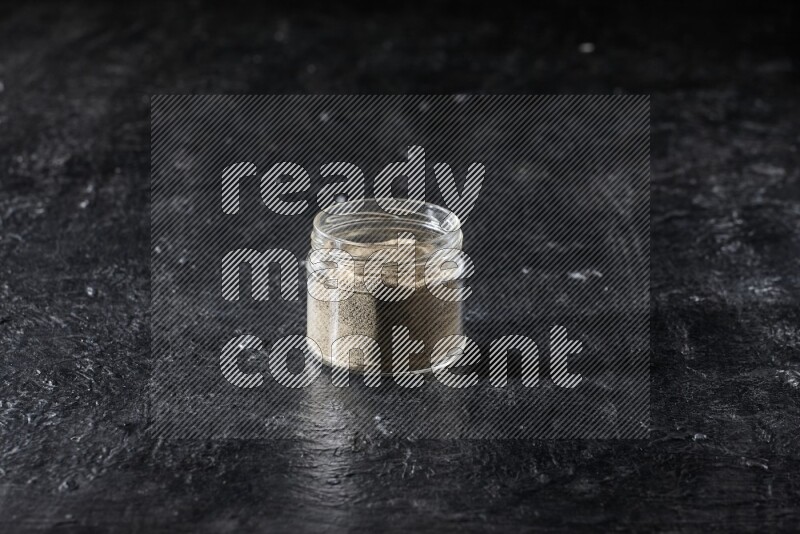 A glass jar full of white pepper powder on textured black flooring