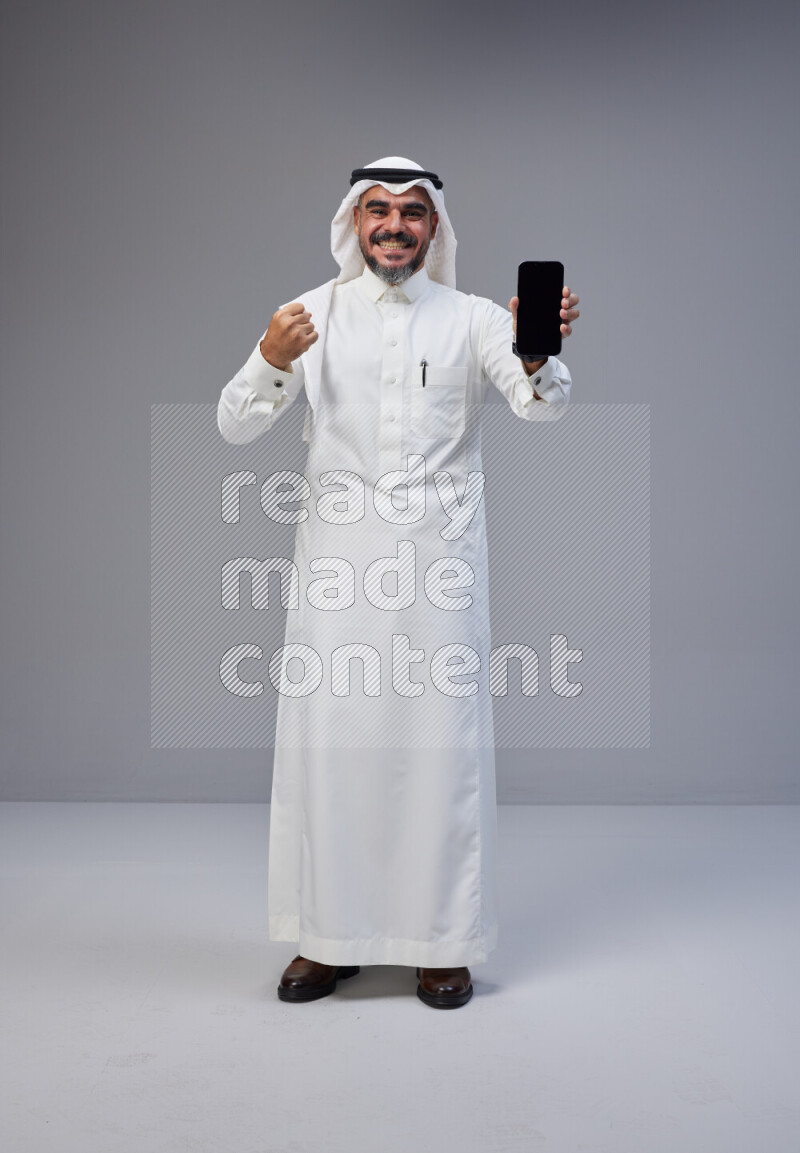 Saudi man Wearing Thob and white Shomag standing showing phone to camera on Gray background
