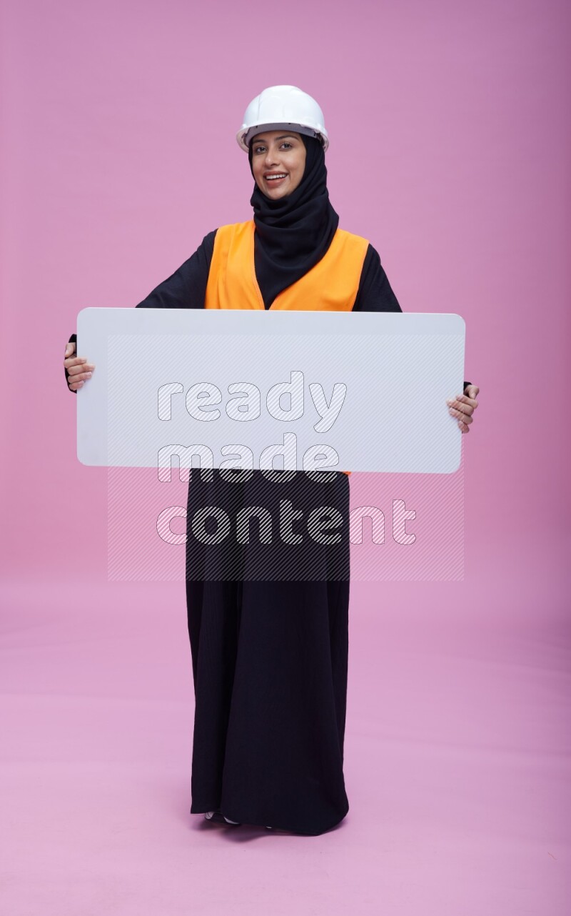 Saudi woman wearing Abaya with engineer vest and helmet standing holding board on pink background