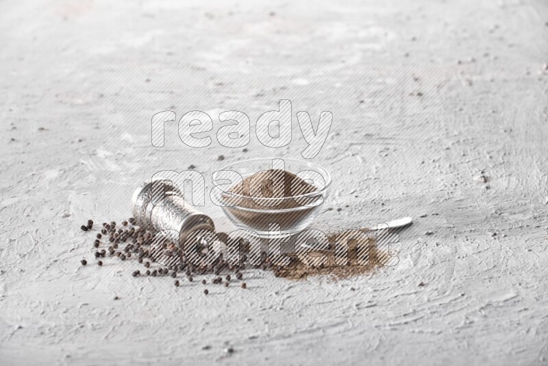 A glass bowl full of black pepper powder with black pepper beads, a turkish metal grinder and a metal spoon on textured white flooring