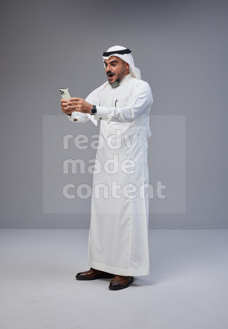 Saudi man Wearing Thob and white Shomag standing texting on phone on Gray background