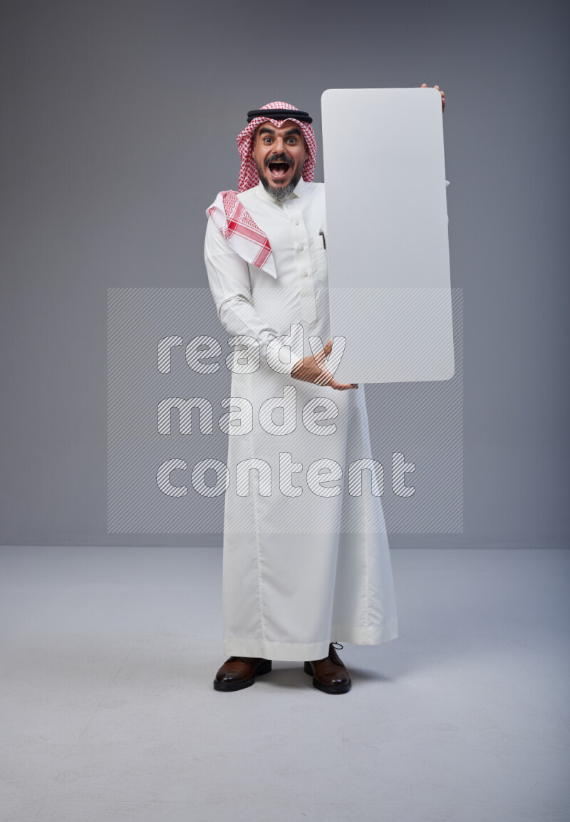 Saudi man Wearing Thob and red Shomag standing holding board on Gray background