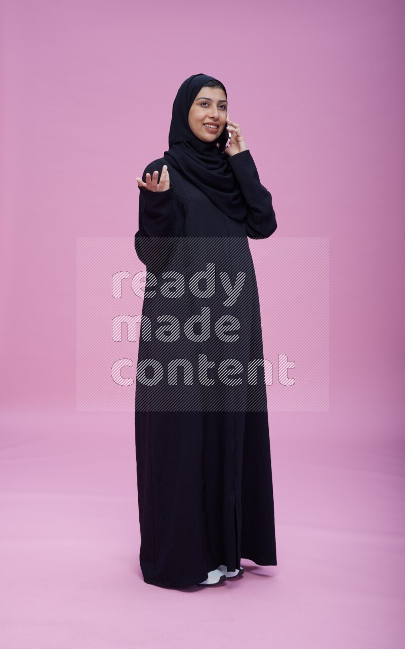 Saudi woman wearing Abaya standing talking on phone on pink background
