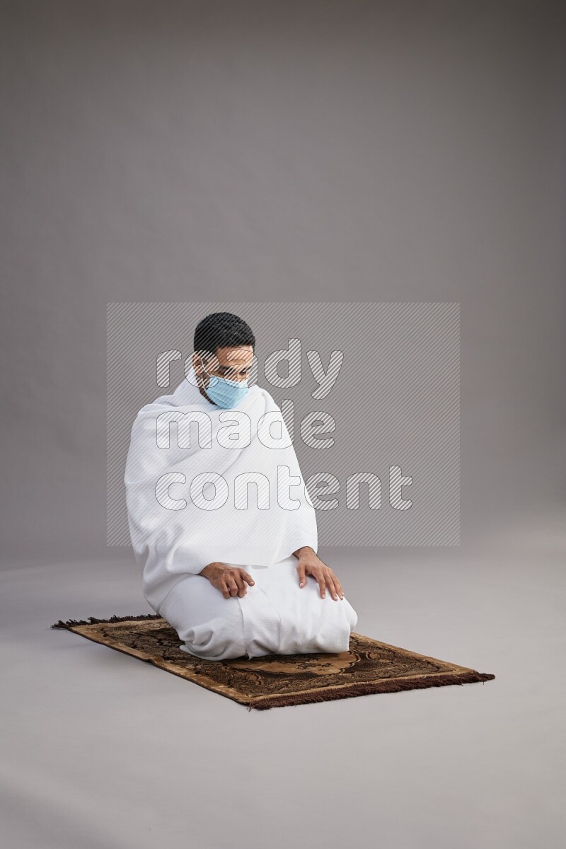 A man wearing Ehram with face mask sitting on floor performing dua'a on gray background