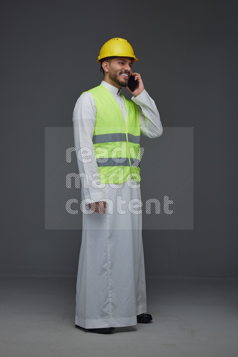 A Saudi man wearing Thobe with a yellow safety vest and white helmet standing and talking in the phone eye level on a gray background