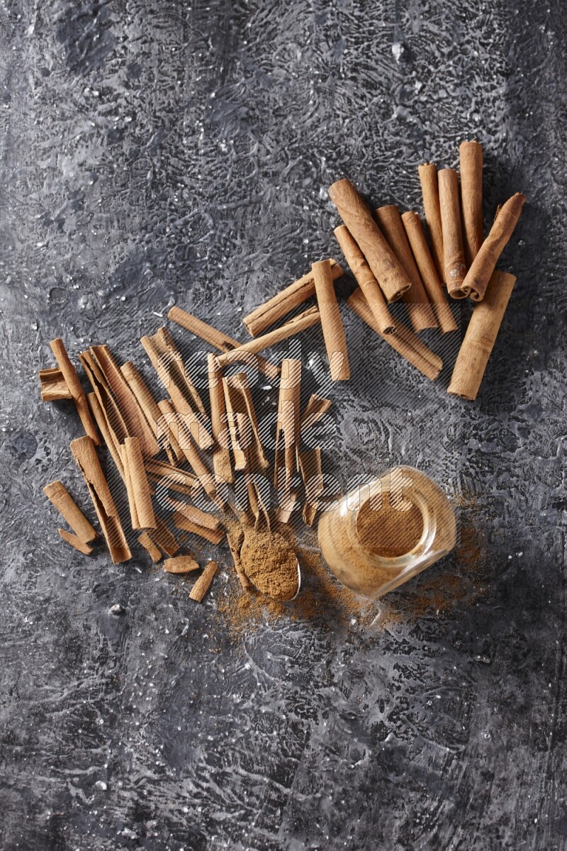 Herbal glass jar and a metal spoon full of cinnamon powder surrounded by cinnamon sticks on textured black background