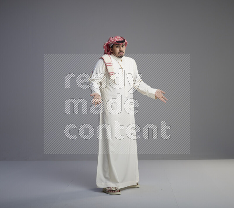 A Saudi man standing wearing thob and red shomag interacting with the camera on gray background