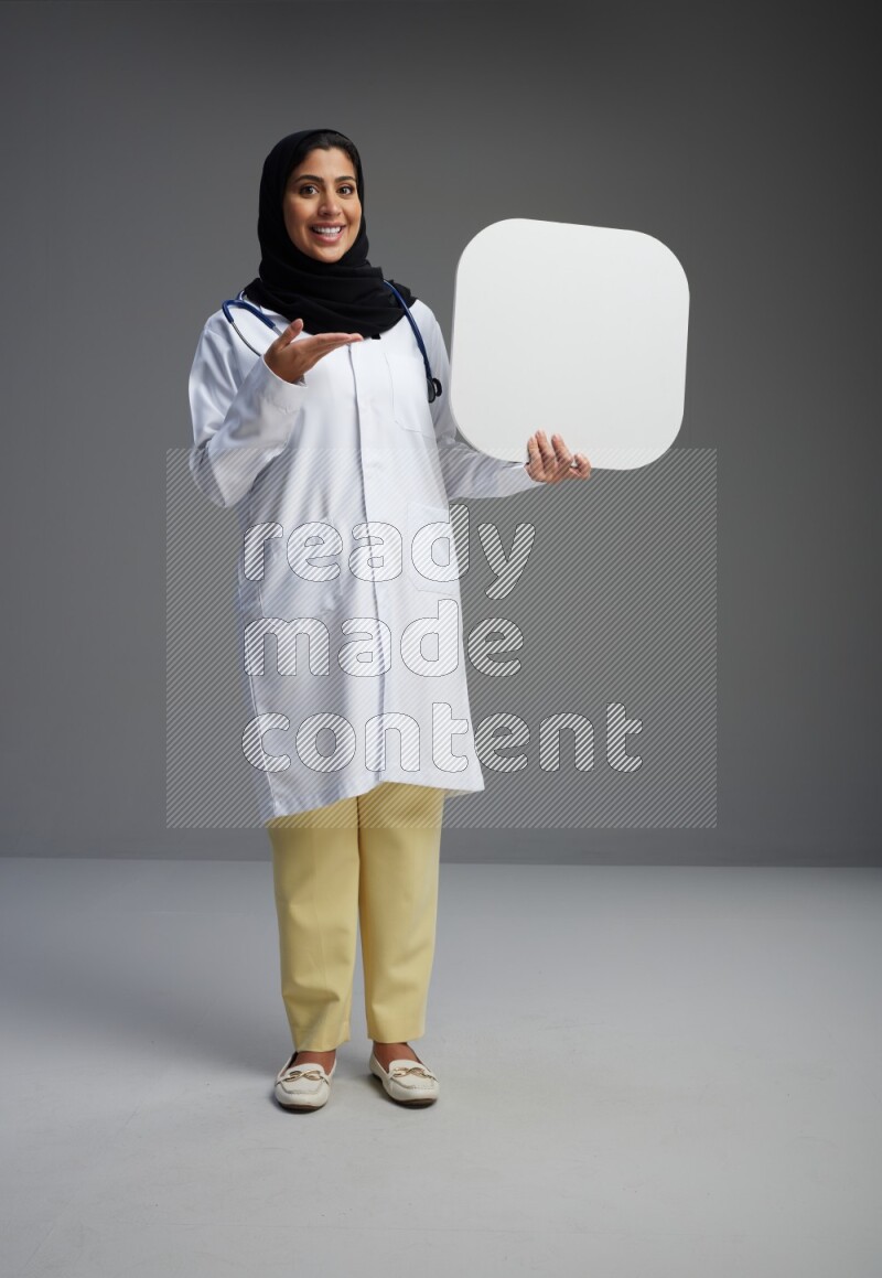 Saudi woman wearing lab coat with stethoscope standing holding social media sign on Gray background